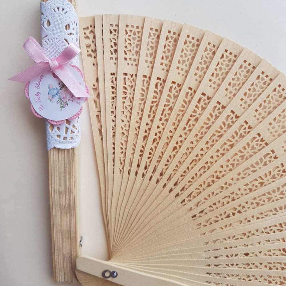Abanicos de madera calados con envoltorio de encaje blanco, moño rosa y etiquetas personalizadas para recuerdos de baby shower.
