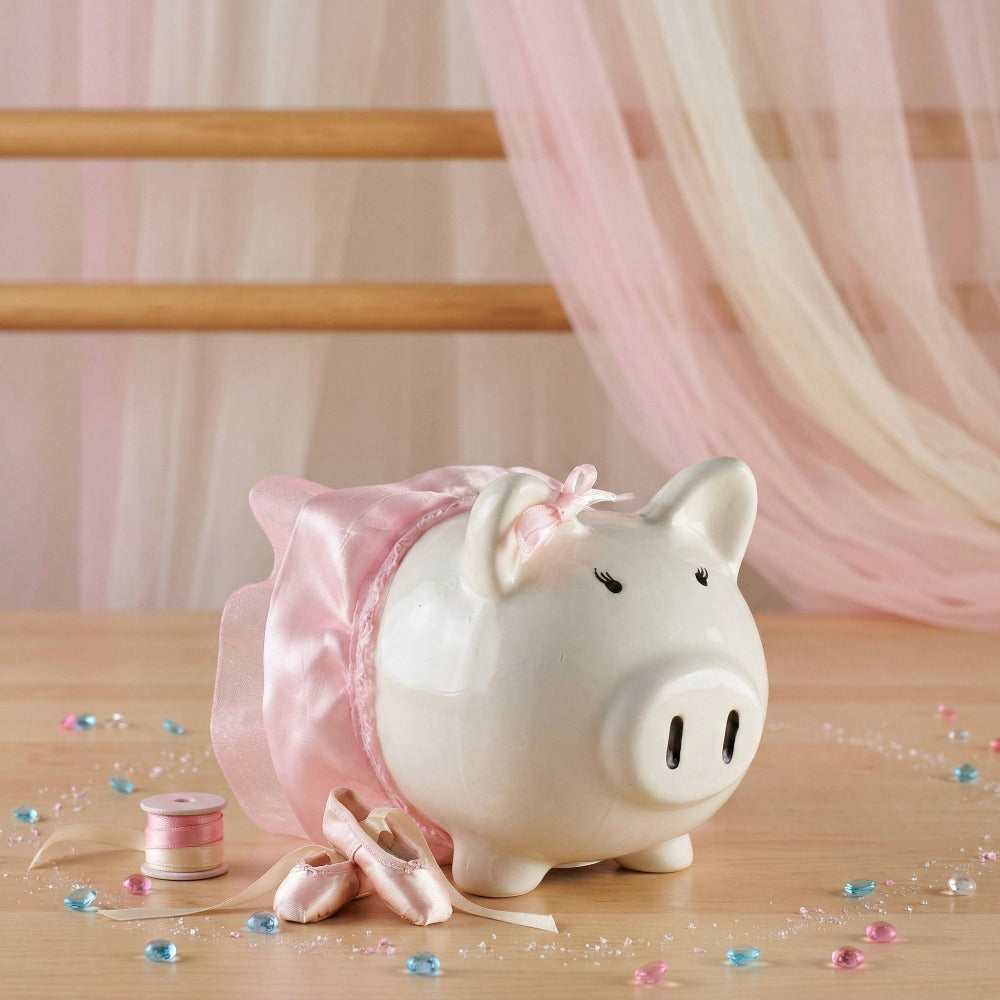 Alcancía de cerdito blanca vestida de bailarina con un tutú rosa, acompañada de zapatillas de ballet en miniatura.