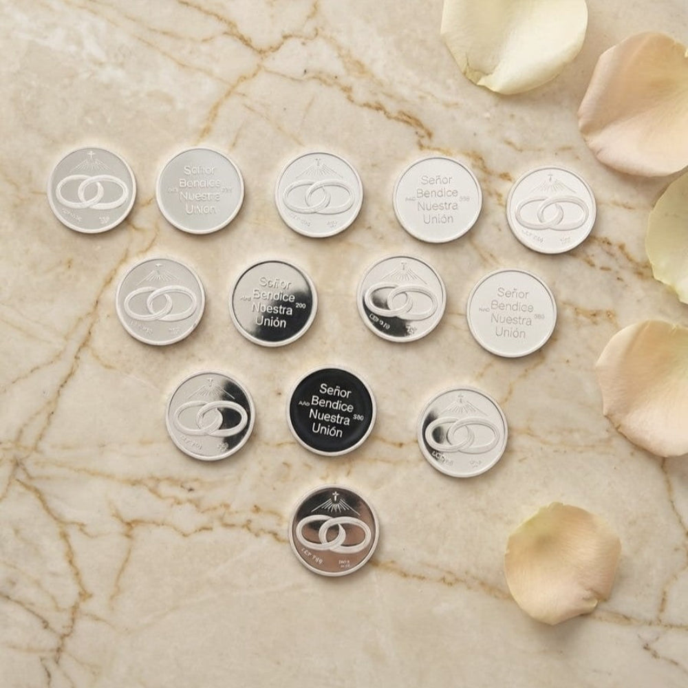 Arras de boda plateadas, con grabados de anillos entrelazados y el texto "Señor Bendice Nuestra Unión", sobre mármol y pétalos de rosa.