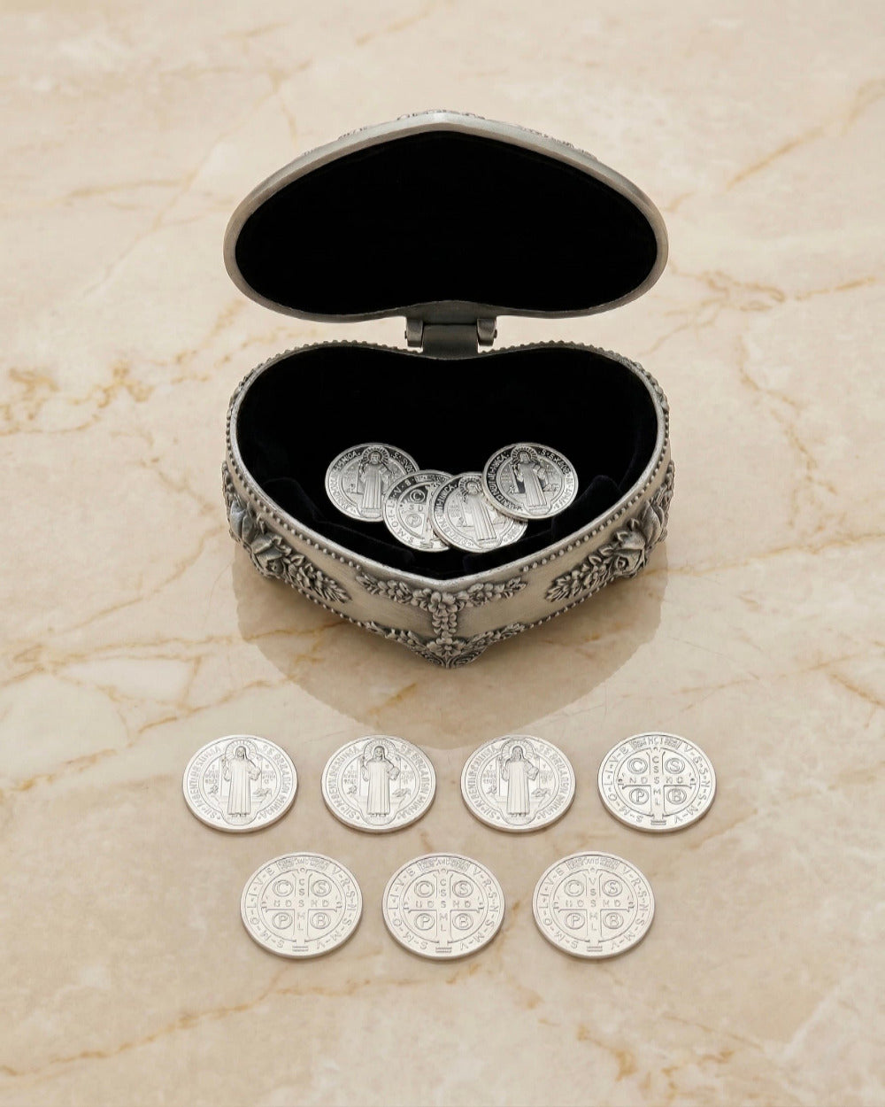 et de 13 arras matrimoniales plateadas (Medallas de San Benito), con cuatro monedas dentro de un estuche de metal plateado ornamentado en forma de corazón abierto con forro de terciopelo negro, y nueve alineadas sobre una superficie de mármol beige pulido.
