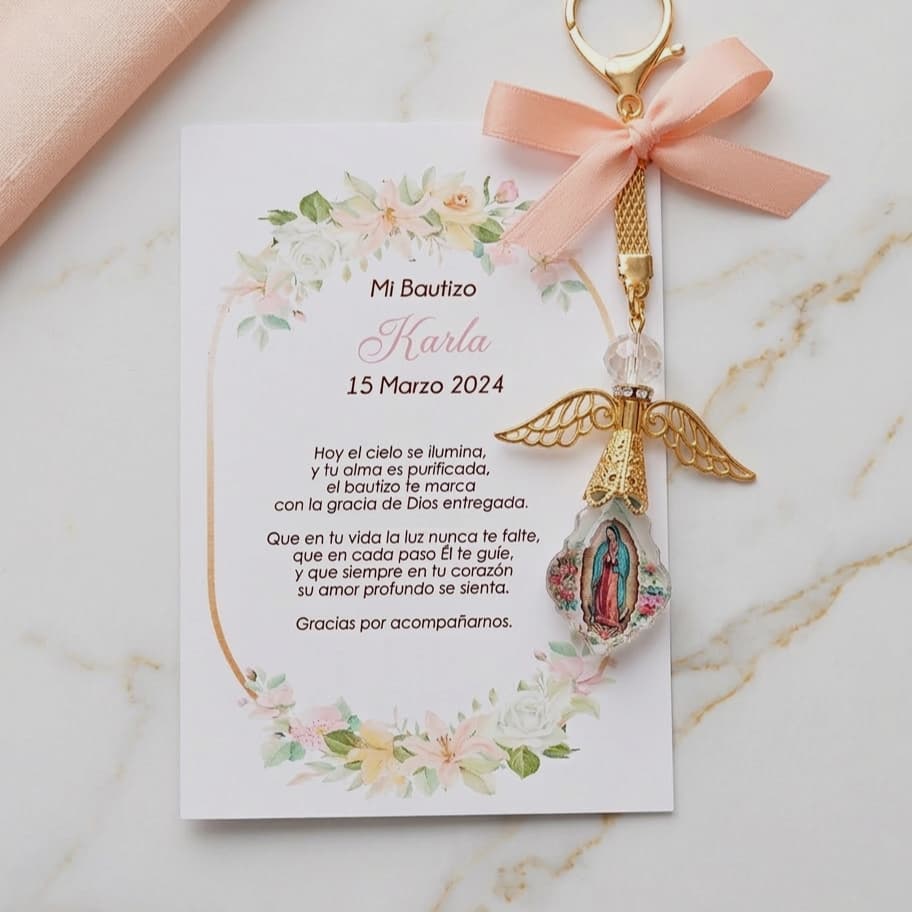 Llavero de angelito dorado con la Virgen de Guadalupe y un moño color durazno, sobre una tarjeta de bautizo decorada con flores tiernas.