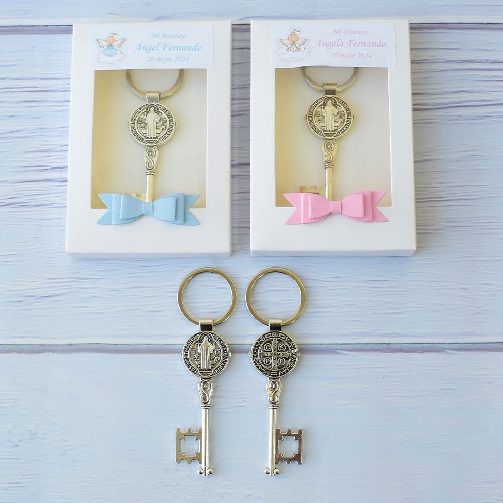Llaveros plateados en forma de llave antigua con la medalla de San Benito. Dos están presentados en cajas blancas como recuerdos de bautizo con pequeños moños en azul y rosa, y otros dos se muestran sueltos en la parte inferior.