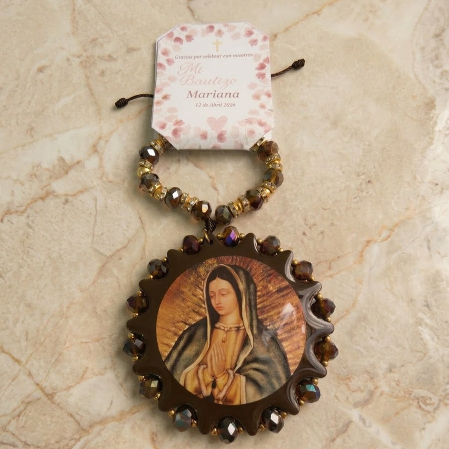 Bendición para coche circular con imagen de la Virgen de Guadalupe, rodeada de cuentas color café oscuro y cristales, colgando de una tira de cuentas a juego con separadores dorados, incluye etiqueta personalizada.