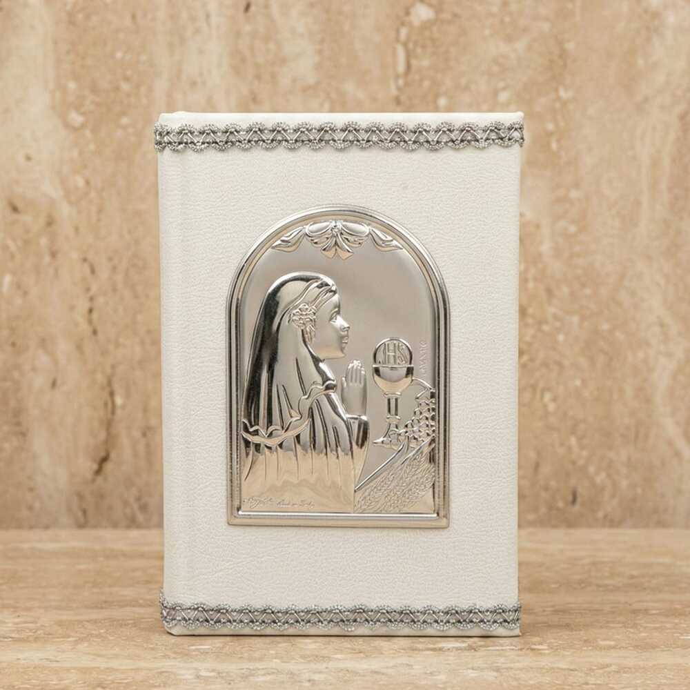 Primer plano de un libro de oraciones blanco con textura sutil. Está decorado con cenefas trenzadas plateadas en los bordes y una placa metálica central en relieve que muestra a una niña rezando, un cáliz y espigas de trigo.