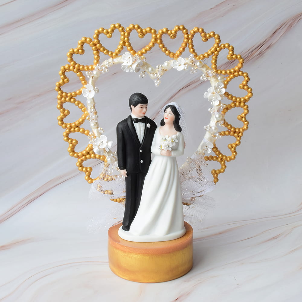 Figura de pastel para boda con novios bajo un arco decorativo en forma de corazón dorado y blanco.