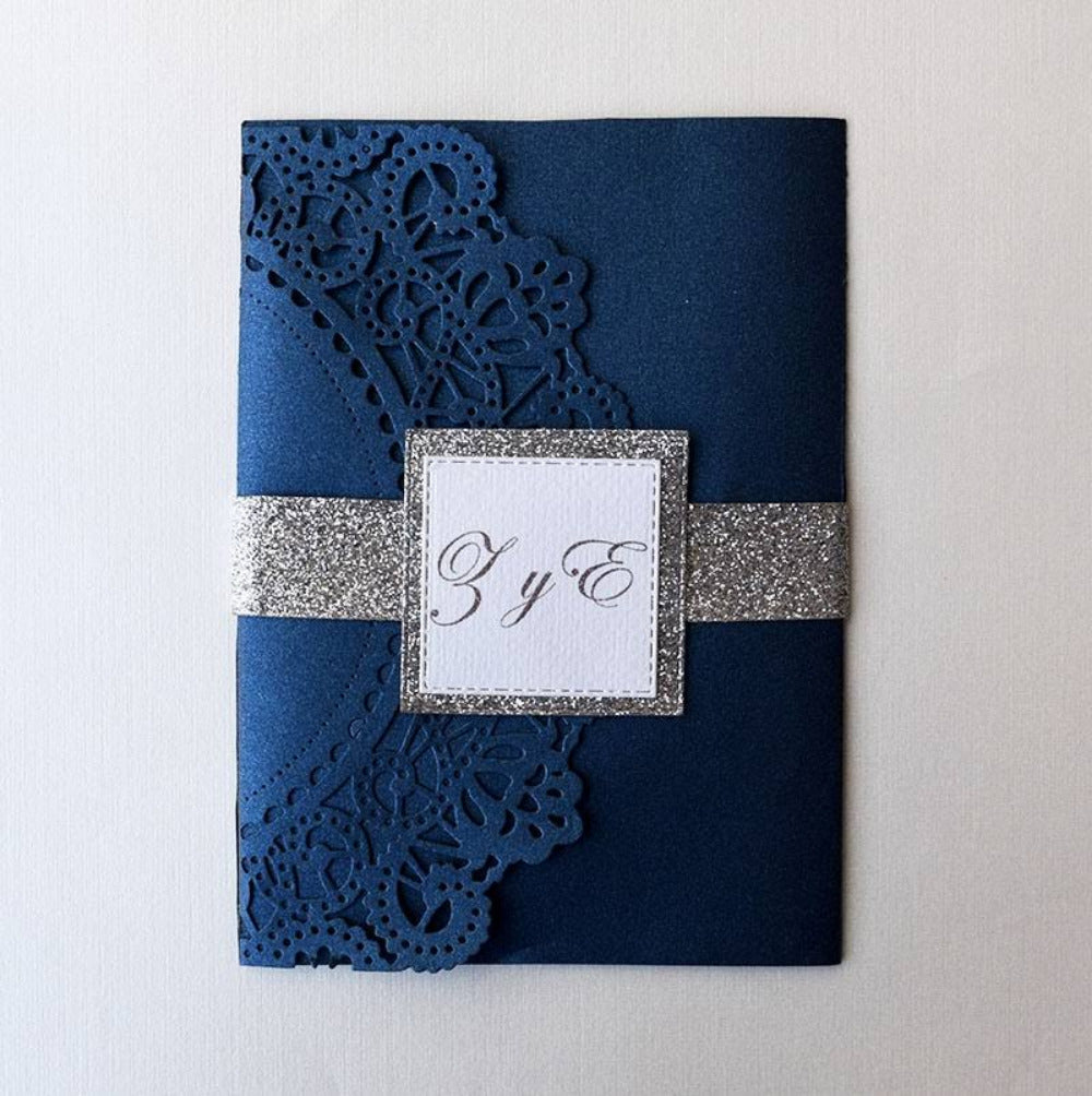 Invitación de boda en color azul marino con elegante corte láser tipo encaje, cintillo de glitter plateado y etiqueta personalizada con iniciales, ideal para eventos nupciales.