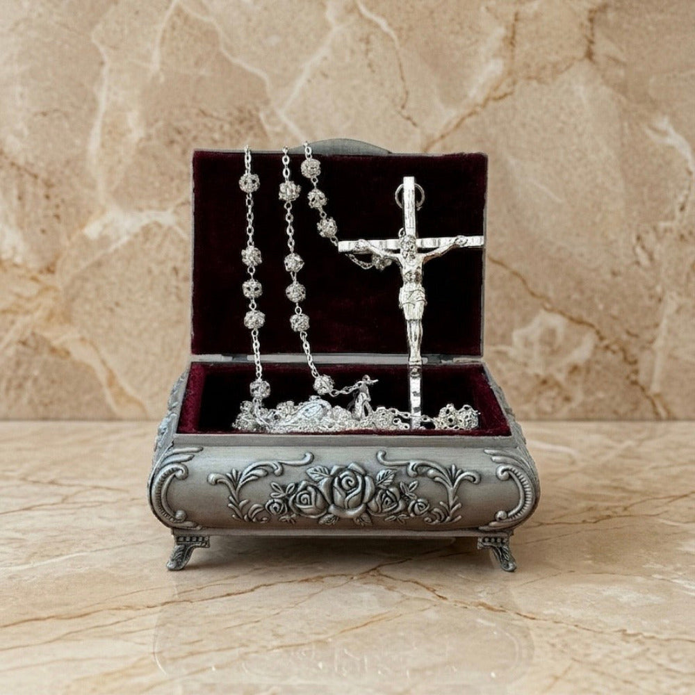 Alhajera metálica plateada con diseño de rosas en relieve, abierta para mostrar un rosario y un crucifijo plateados con Cristo, forrada en terciopelo color burdeos. Ideal como recuerdo de boda o evento religioso.