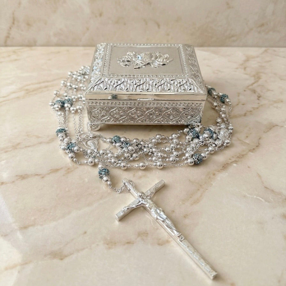Lazo de boda plateado con crucifijo y cuentas decoradas, acompañado de un cofre metálico con relieves.