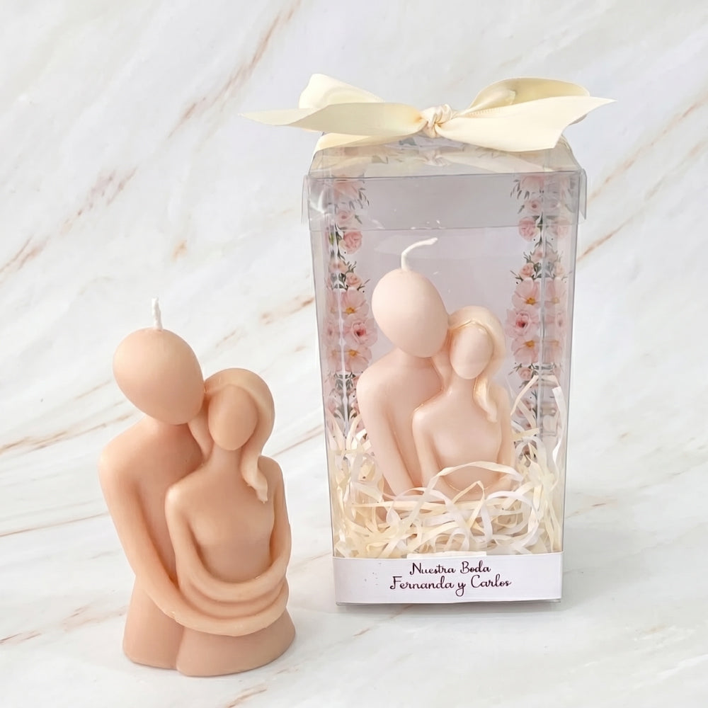 Dos velas beige con forma de pareja abrazada sobre mármol, una en caja de regalo transparente con un lazo.