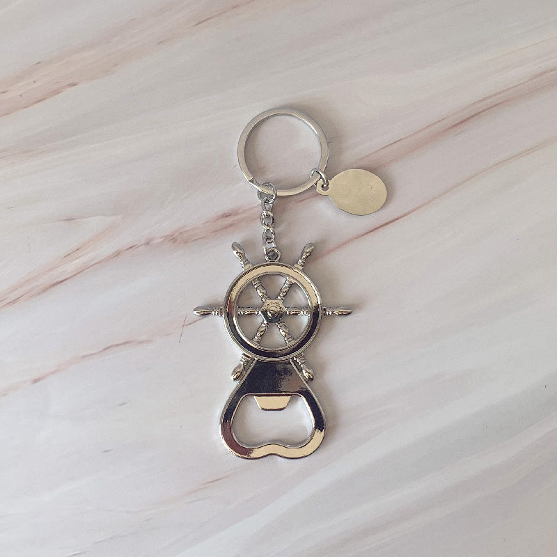 Llavero destapador metálico con diseño de timón náutico, ideal para recuerdos de eventos sociales, sobre fondo de mármol.