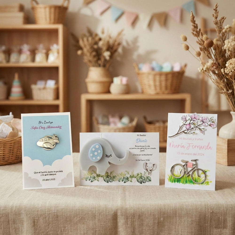 Tres recuerdos de bautizo personalizados sobre una mesa beige: una tarjeta azul con dije de avión, una vela de elefante gris y una tarjeta floral con dije de bicicleta.