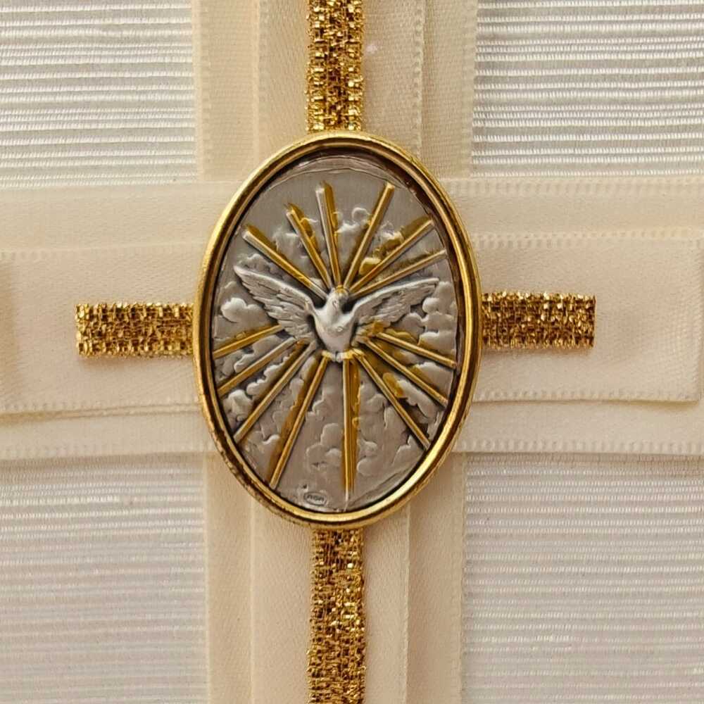 Primer plano de un medallón religioso ovalado con una paloma blanca y rayos dorados, montado sobre una cruz de tela marfil con detalles de cinta dorada brillante.