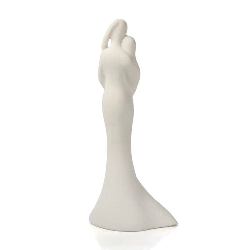 Figura decorativa de novios abrazados en cerámica blanca con detalle de corazón y calados sobre fondo rosa marmoleado.