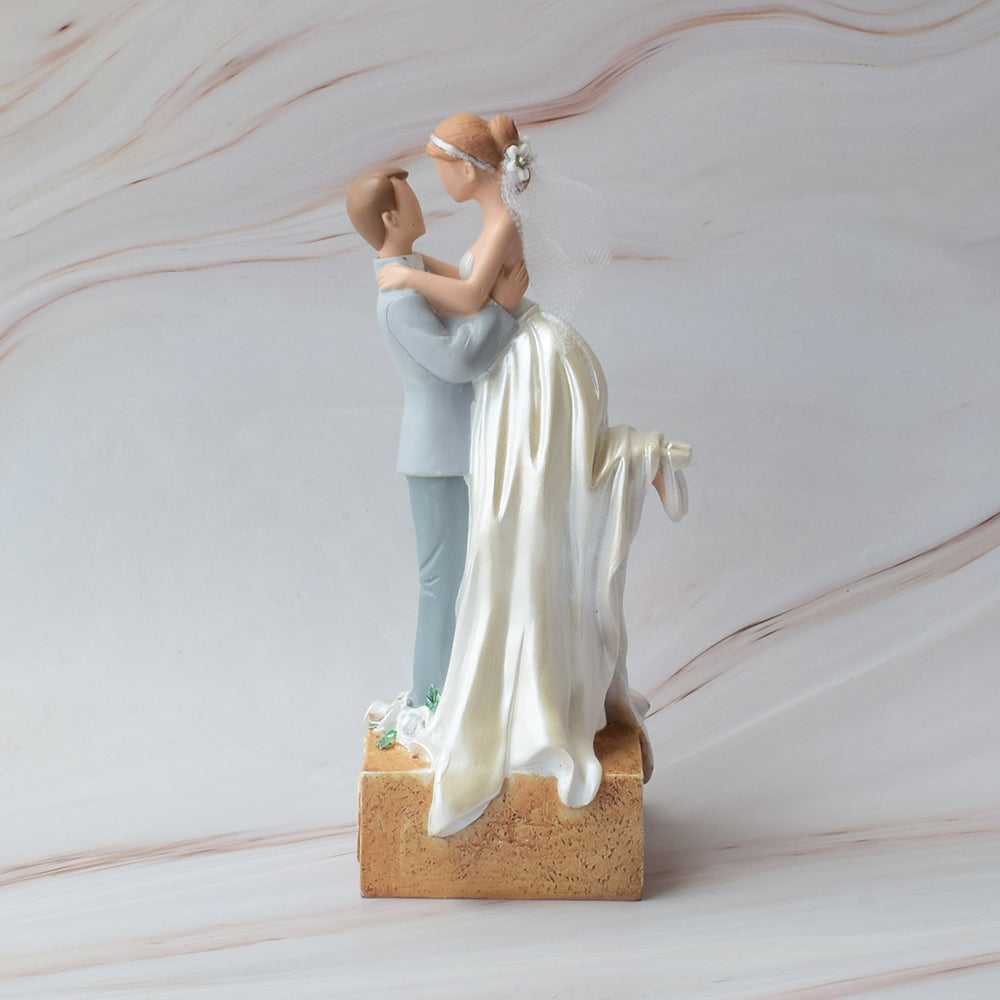 Figura para pastel de boda con novios abrazados, él cargándola, con acabados brillantes y base decorativa sobre mármol.