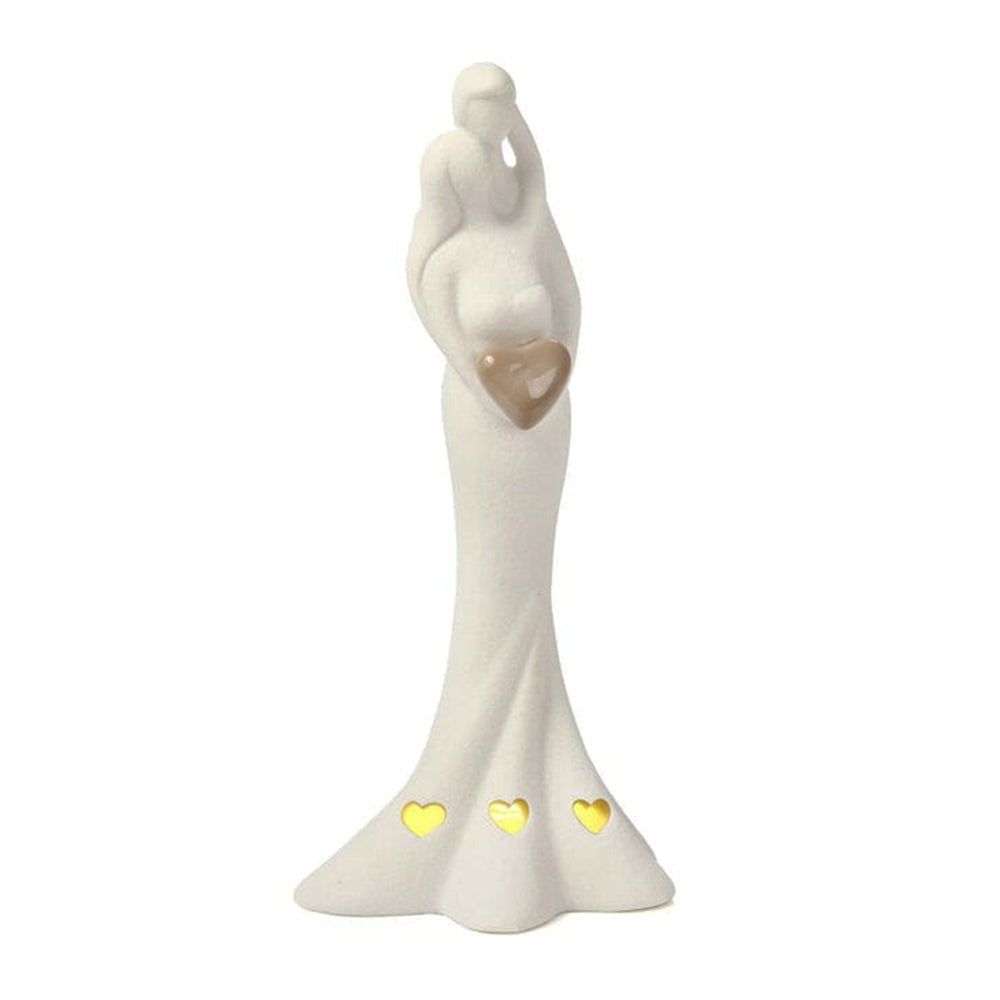Figura decorativa de novios abrazados en cerámica blanca con detalle de corazón y calados sobre fondo rosa marmoleado.