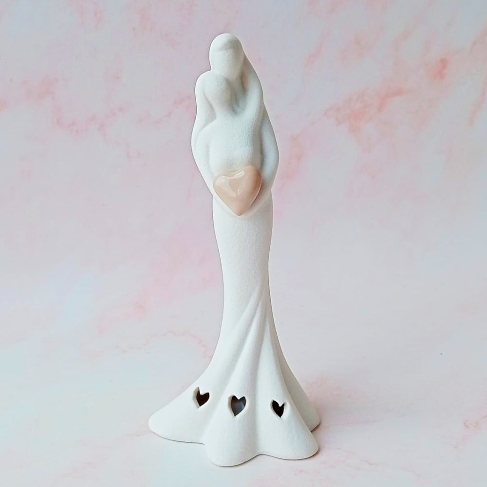 Figura decorativa de novios abrazados en cerámica blanca con detalle de corazón y calados sobre fondo rosa marmoleado.