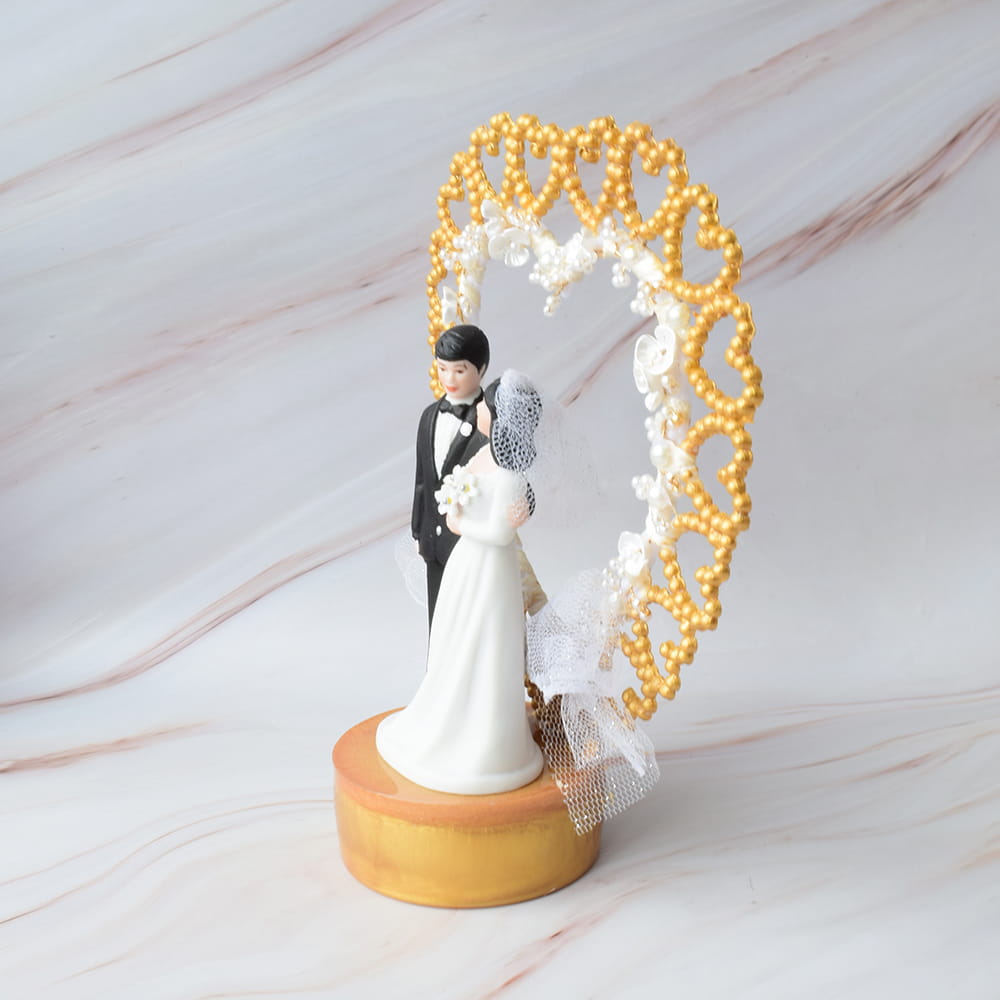 Figura para pastel de boda con pareja de novios, base circular y arco decorativo en forma de corazón con cuentas doradas y tul.
