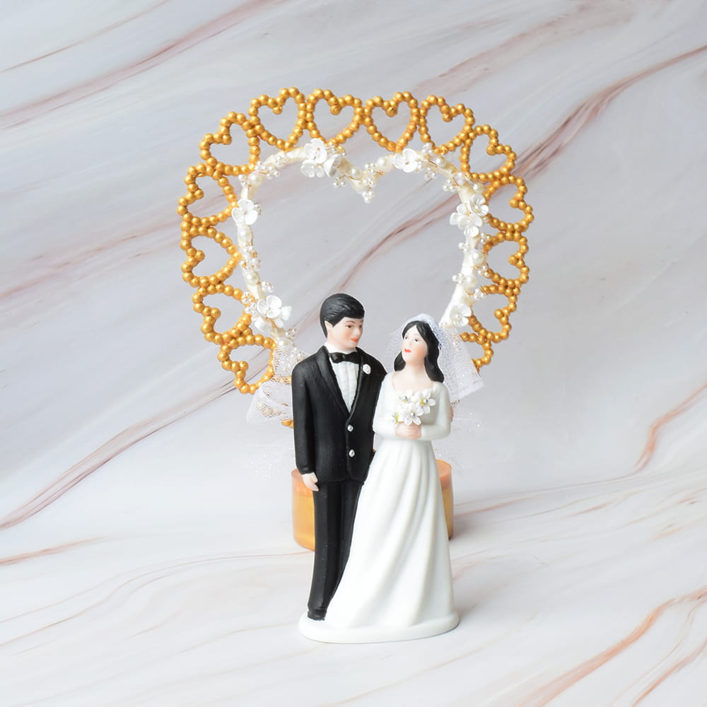 Figura para pastel de boda con pareja de novios y respaldo en forma de corazón dorado con detalles de perlas y flores blancas, ideal para celebración en CDMX.