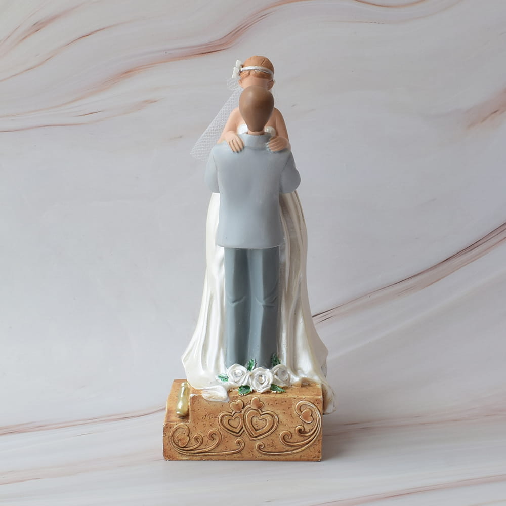Muñecos para pastel de boda vistos desde atrás, novio cargando a la novia con detalle de rosas blancas en la base.