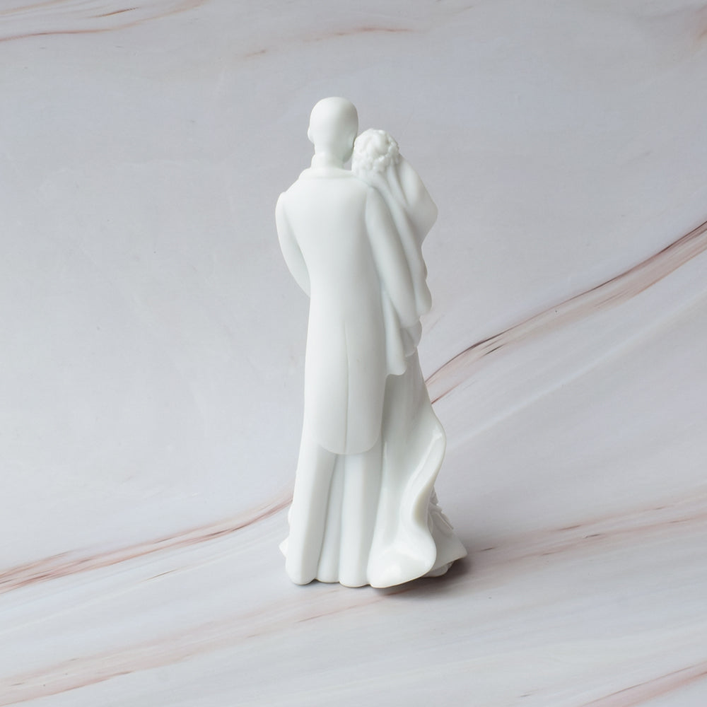 Figura para pastel de boda en cerámica blanca con novios abrazados y detalles florales en relieve sobre mármol.