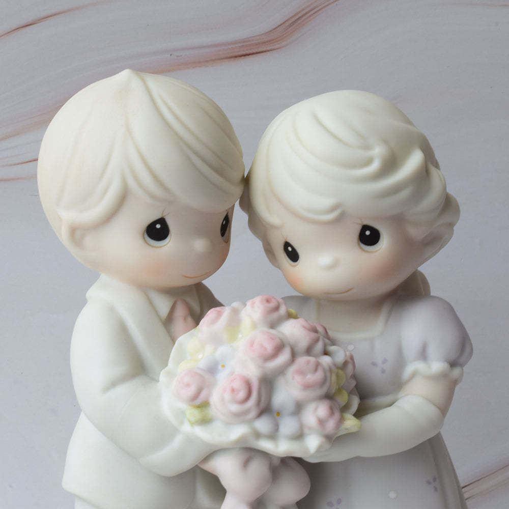 Figura para pastel de boda en cerámica con detalles de flores y la frase "Love Vows To Always Bloom", sobre fondo de mármol.
