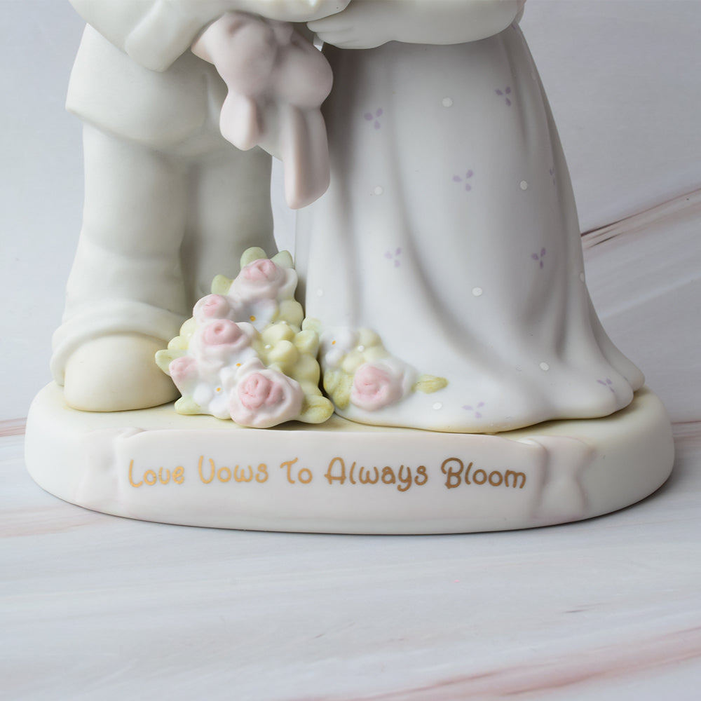 Figura para pastel de boda en cerámica con detalles de flores y la frase "Love Vows To Always Bloom", sobre fondo de mármol.