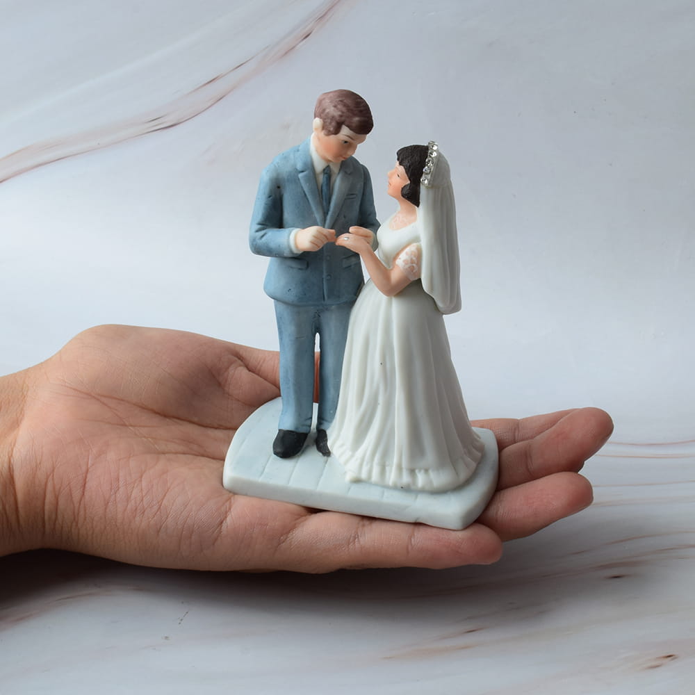 Figura pequeña para pastel de boda sostenida sobre una mano, con el novio en traje azul claro colocando un anillo a la novia en vestido blanco.