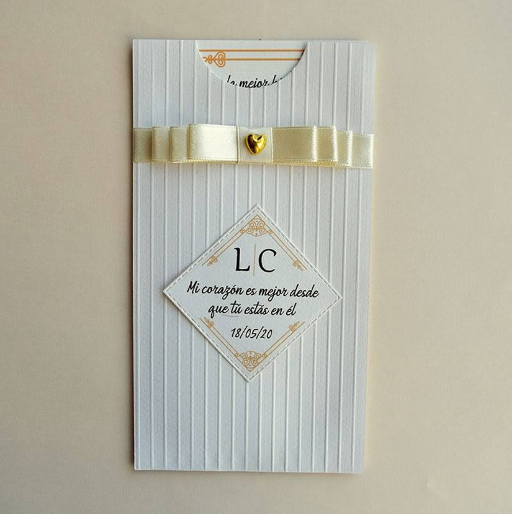 Invitación de boda con sobre blanco de textura lineal, decorada con un cintillo de satén en color crema, dije de corazón dorado y etiqueta romántica personalizada, ideal para eventos nupciales.