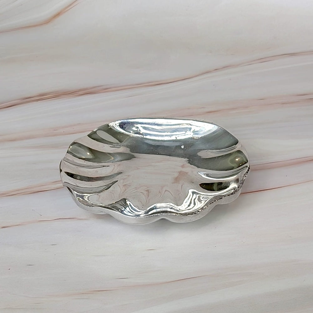 Concha de bautismo de pewter pulida con bordes ondulados sobre superficie de mármol.