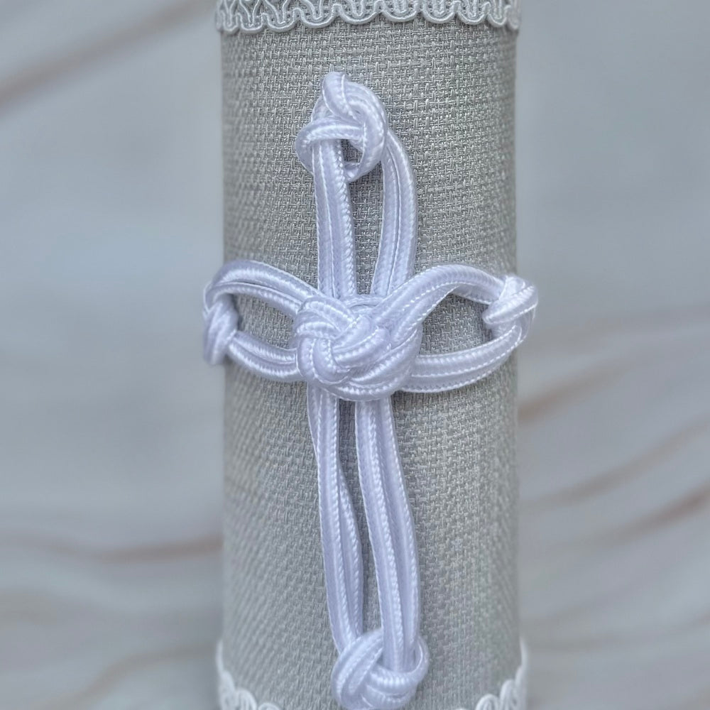 Cirio de bautizo con cruz artesanal de cordón blanco satinado sobre tela de lino gris con pasamanería decorativa.