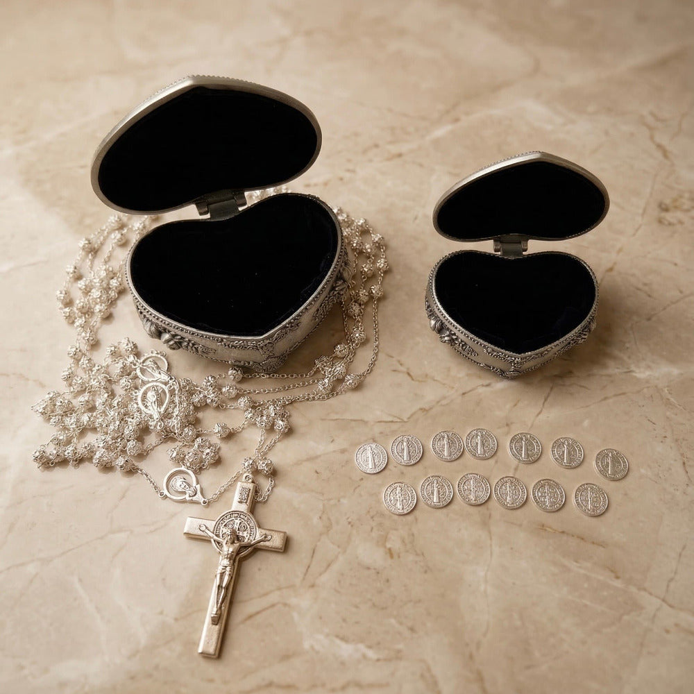 Set de lazo de boda y 13 arras matrimoniales plateadas, junto a dos estuches metálicos en forma de corazón abiertos mostrando su interior oscuro, sobre superficie de mármol.