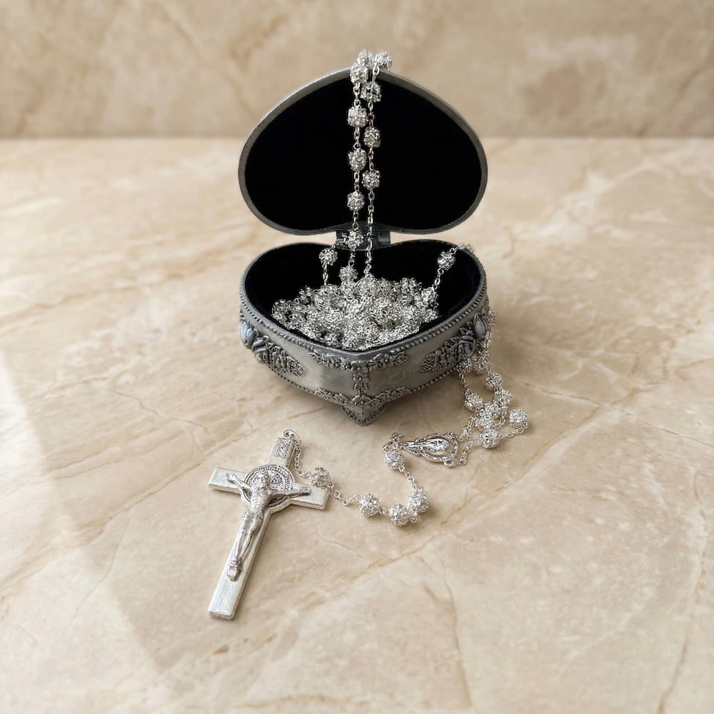 Lazo de plata con cuentas texturizadas y crucifijo a juego, presentado en un estuche metálico ornamentado en forma de corazón abierto con forro de terciopelo negro, sobre una superficie de mármol beige.