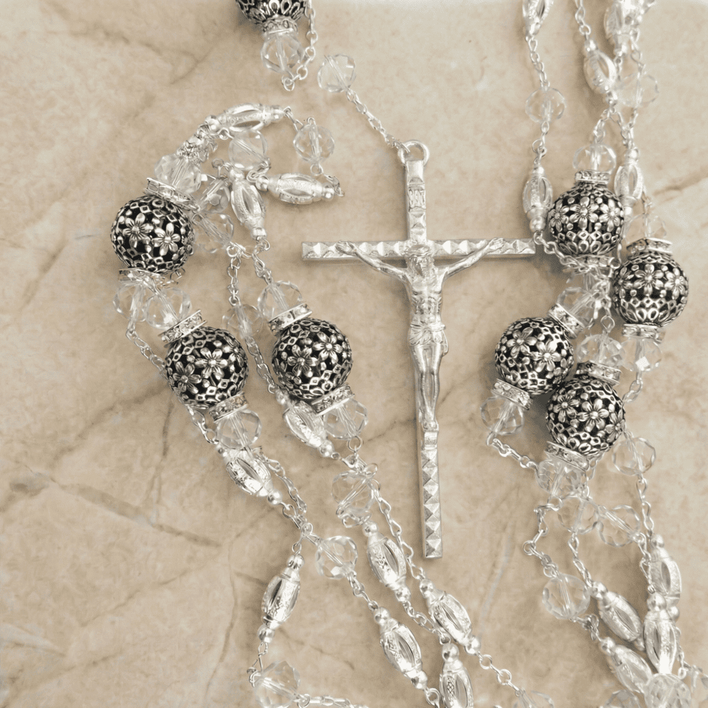 Lazo de boda con crucifijo plateado, cuentas de cristal translúcido y esferas oscuras con detalles florales calados en plata.