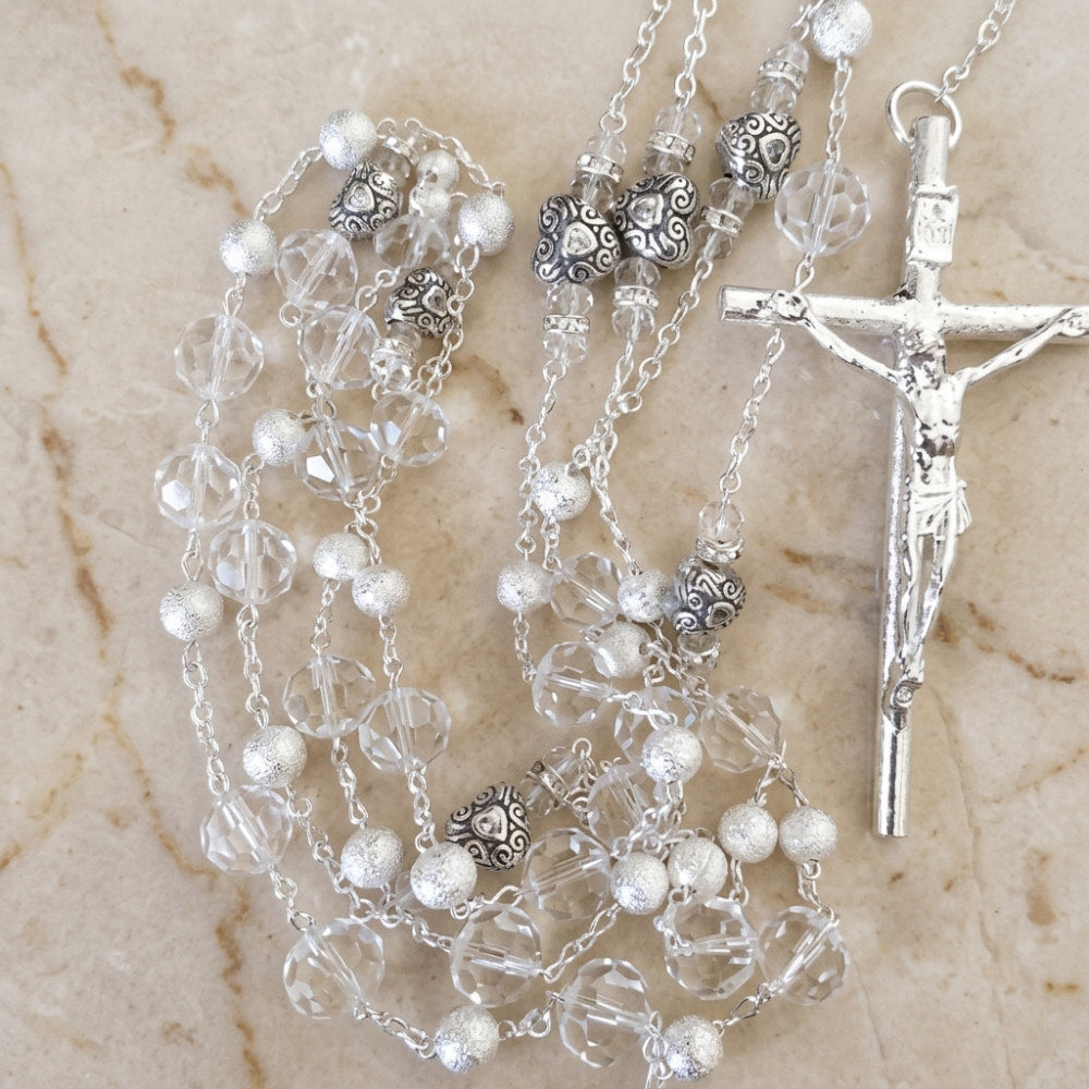 Lazo de boda en baño de plata con cuentas de cristal y corazones, sobre superficie de mármol beige.