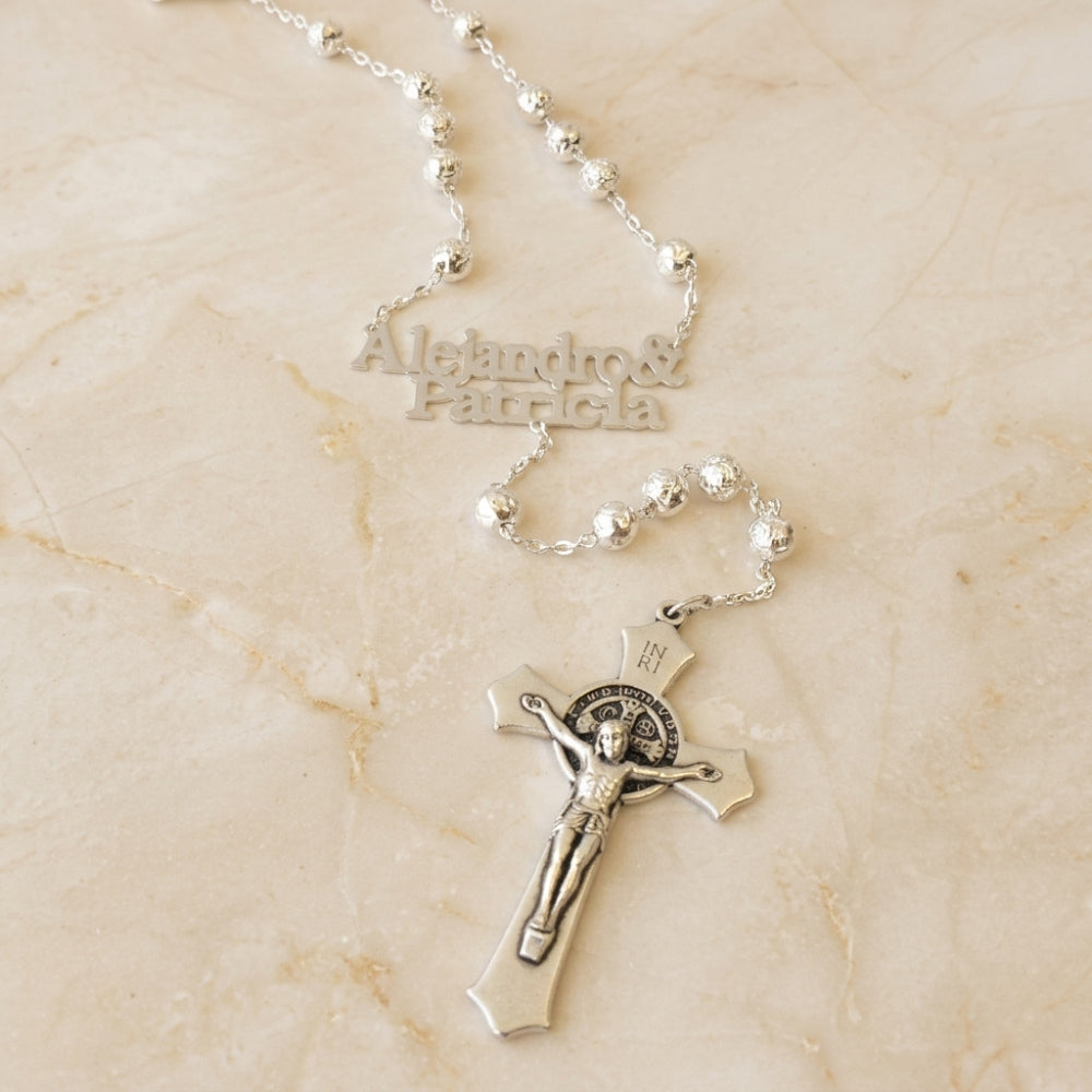 Rosario de plata personalizado con nombres 'Alejandro & Patricia' y cruz de San Benito, sobre mármol.