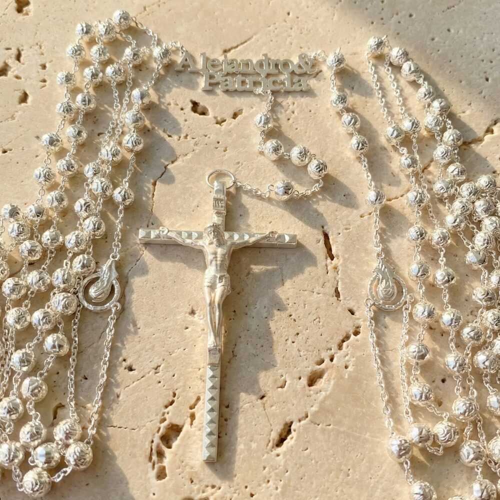 Lazo matrimonial de plata en forma de rosario sobre una superficie de piedra clara. Destaca un crucifijo central detallado, cuentas redondas texturizadas y los nombres 'Alejandro & Patricia' integrados en la parte superior de la cadena metálica.