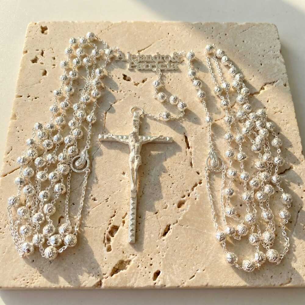 Lazo de boda plateado personalizado con crucifijo, cuentas de la Virgen, con los nombres "Alejandro & Patricia", sobre piedra travertino beige.