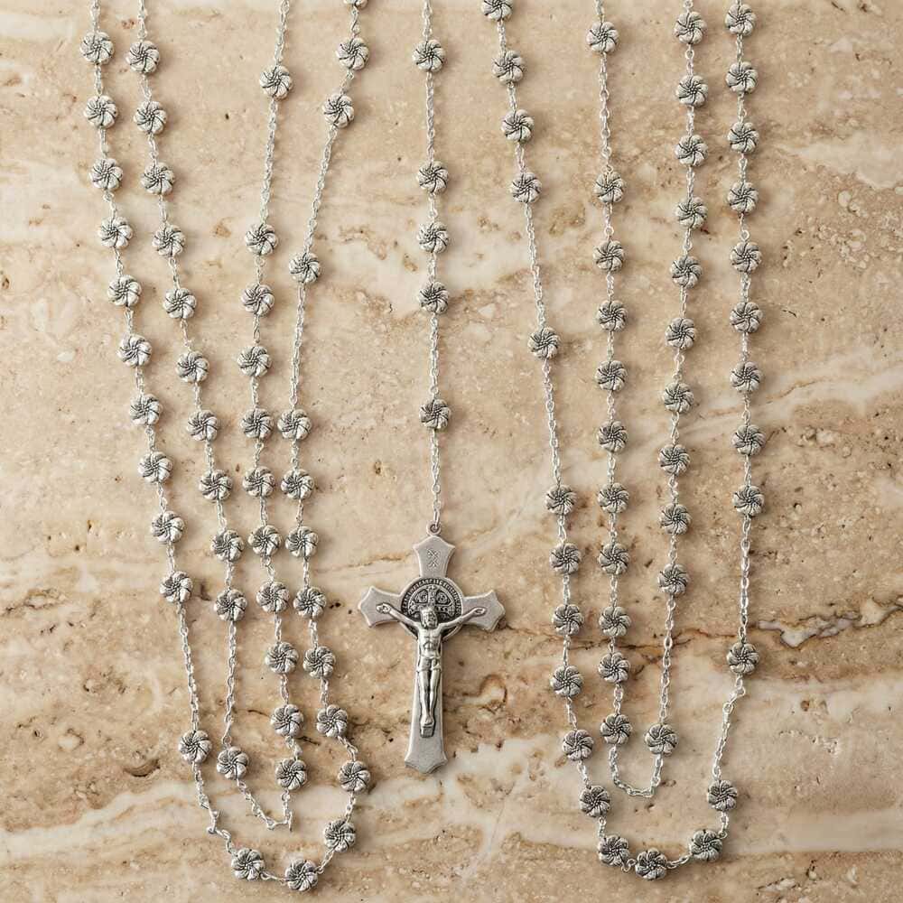 lazo para boda católica de plata, cuentas con forma de flor texturizada y un crucifijo de San Benito, sobre una superficie de mármol beige pulido.