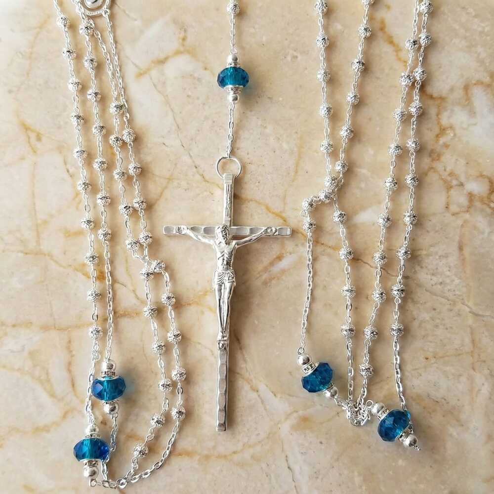 Lazo para boda de plata con cuentas texturizadas, detalles de cristales azul turquesa y crucifijo central sobre mármol beige.