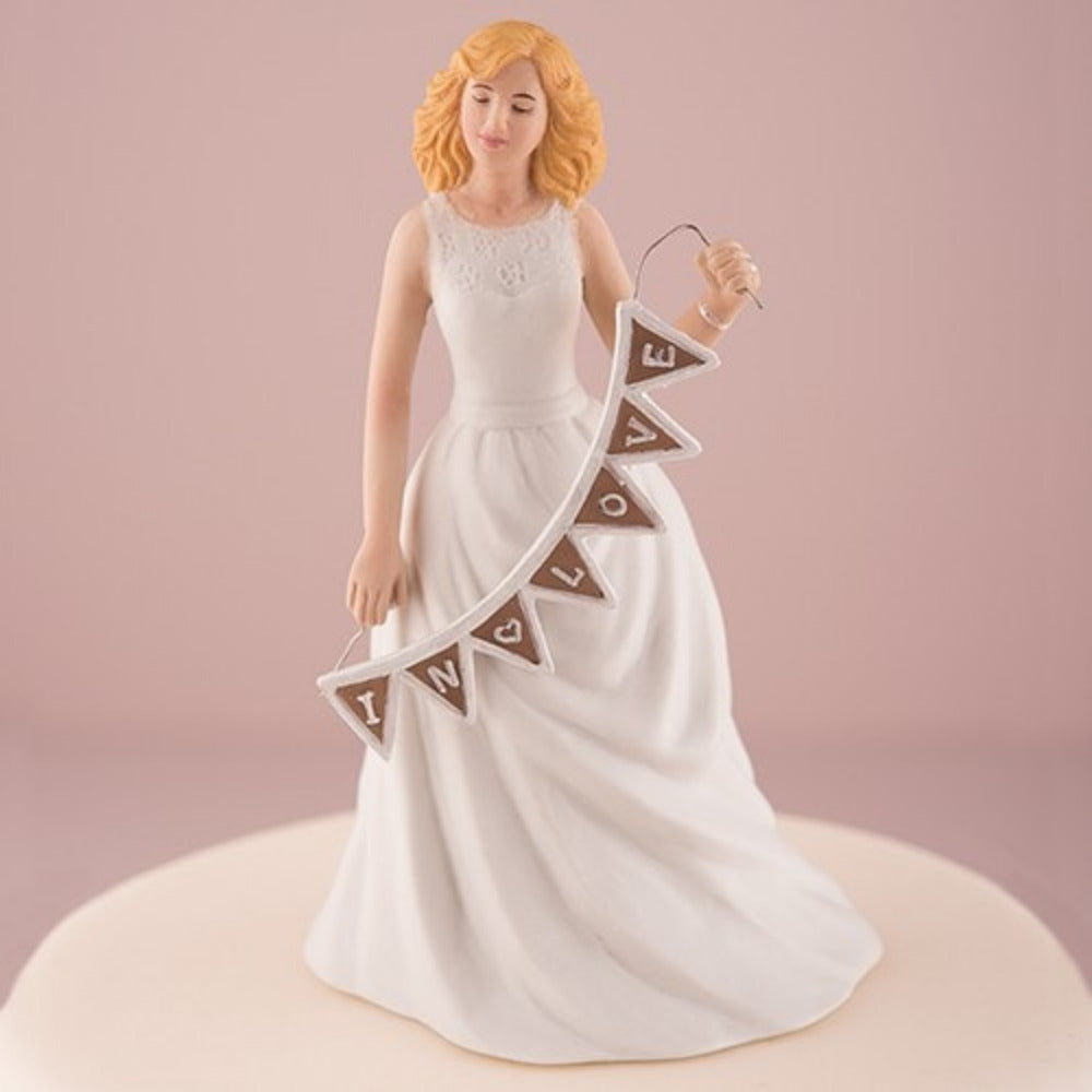 Muñeco para pastel de boda con figura de novia rubia sosteniendo un banderín con la frase "In Love", ideal para decoración de pasteles de nupcias y eventos sociales de Ondine Collection.