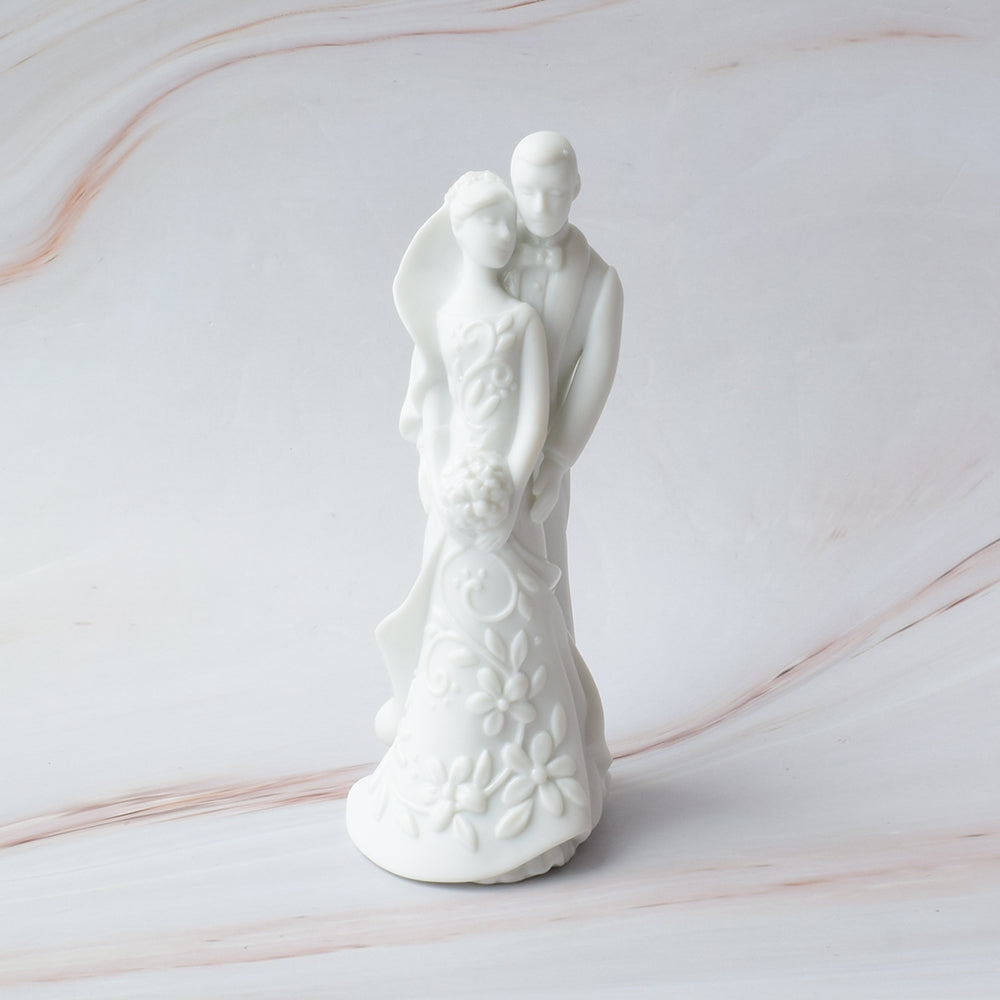 Figura para pastel de boda en cerámica blanca con novios abrazados y detalles florales en relieve sobre mármol.
