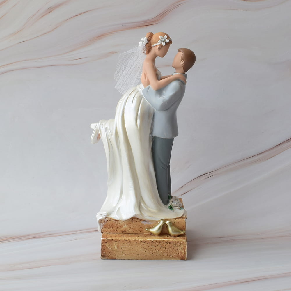 Figura para pastel de boda que muestra al novio cargando a la novia en brazos, sobre una base rústica con dos pájaros dorados.