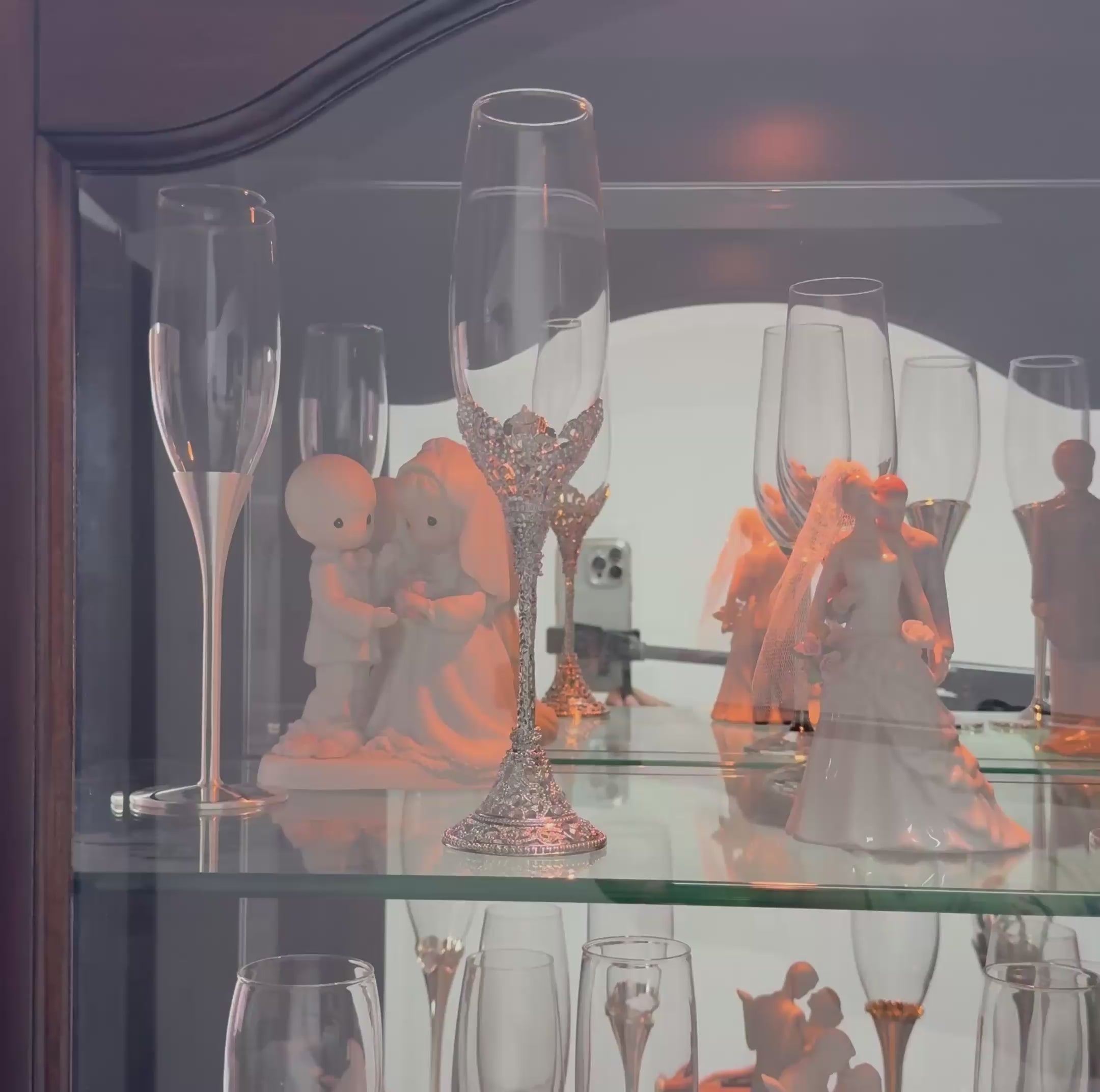 Cargar vídeo: Colección de accesorios para matrimonio en exhibición, destacando copas de cristal decoradas, figuras decorativas de novios y cristalería fina para eventos nupciales, presentada dentro de una vitrina de cristal iluminada.