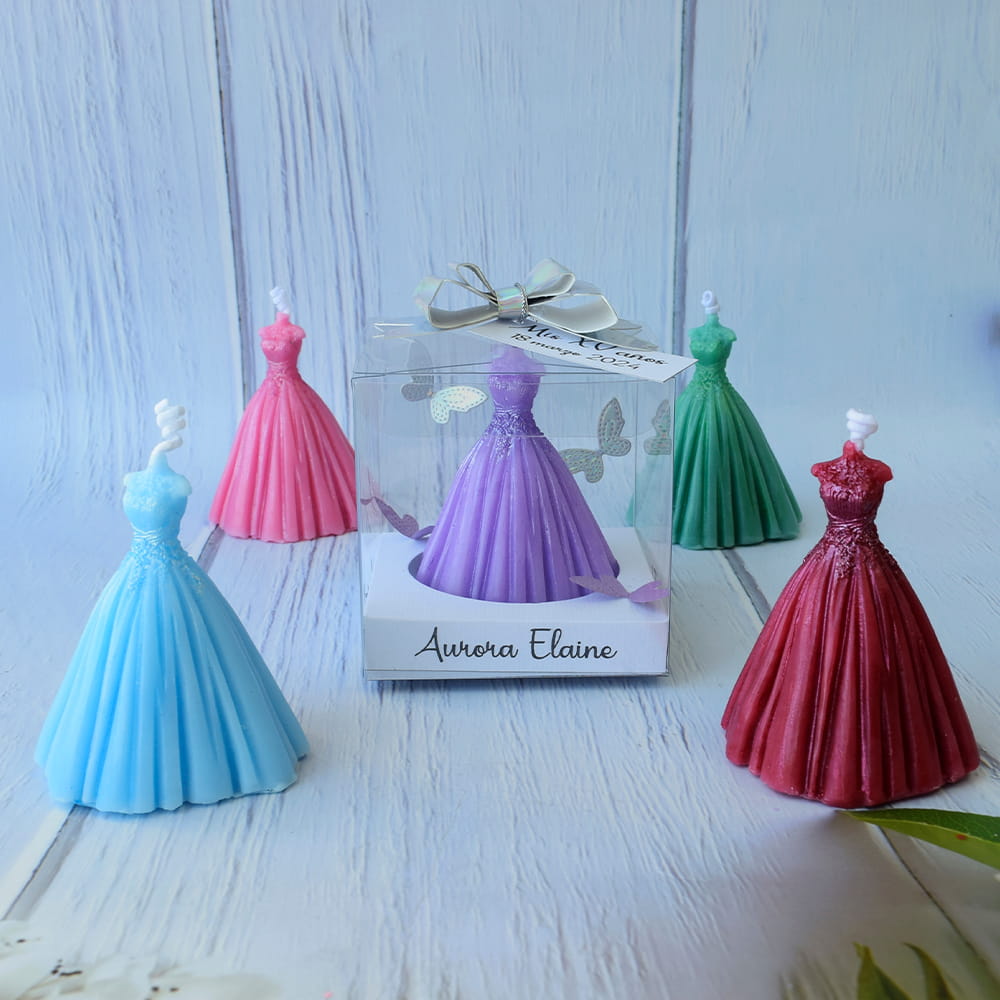 Recuerdo original para quinceañera. Velas decorativas en forma de vestido de gala disponibles en varios colores. Se muestra la presentación final en caja transparente con etiqueta personalizada y mariposas decorativas, ideal como detalle para los invitados del evento.