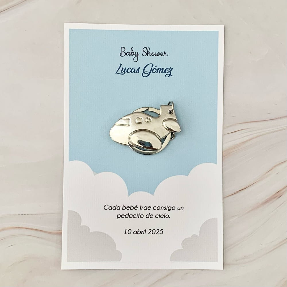Llavero plateado de avioncito sobre tarjeta azul con nubes y texto personalizado, ideal para recuerdo de baby shower de niño.