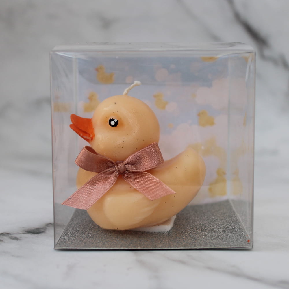 Vela artesanal en forma de patito amarillo con moño de listón color malva, recuerdo para baby shower en caja de acetato con fondo decorativo de patitos.