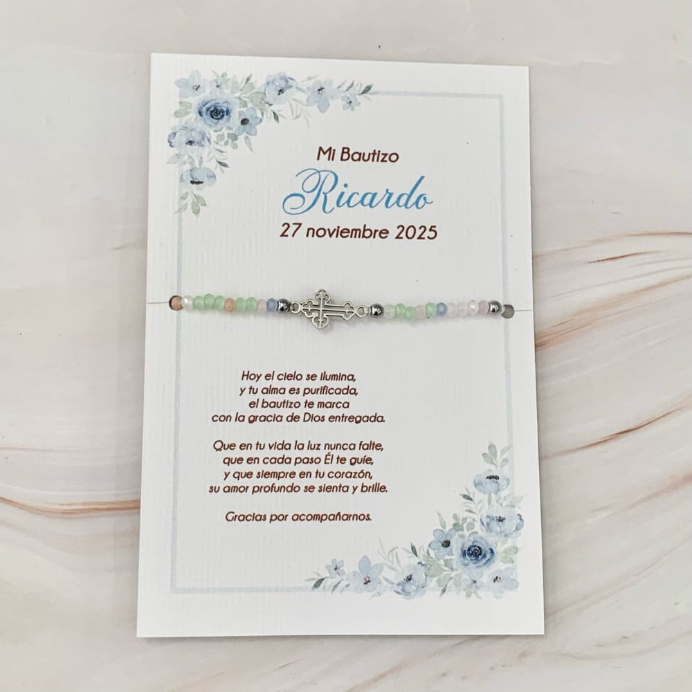 Pulsera con dije de cruz metálica sobre tarjeta personalizable de recuerdo para bautizo con flores azules de acuarela.