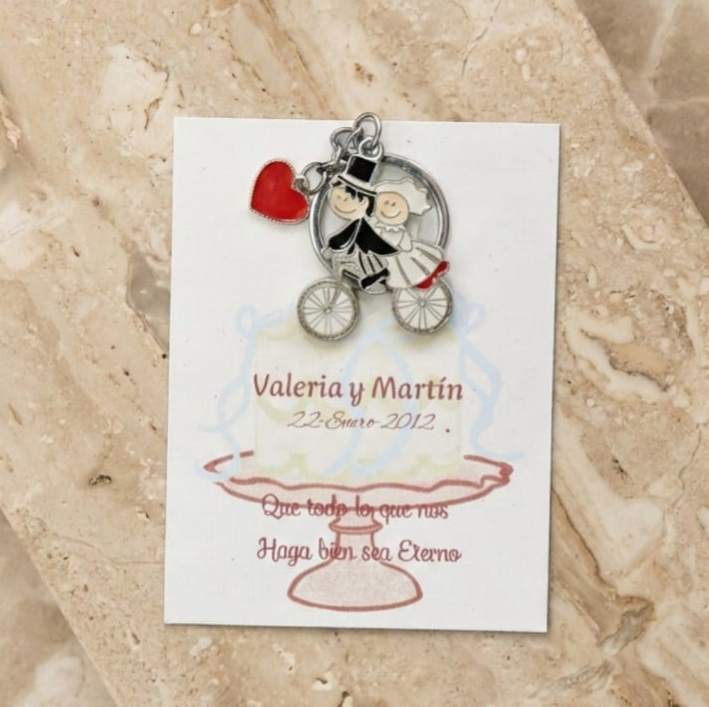 Recuerdo de boda con llavero de novios en bicicleta y dije de corazón rojo, montado en tarjeta personalizada con nombres y fecha.