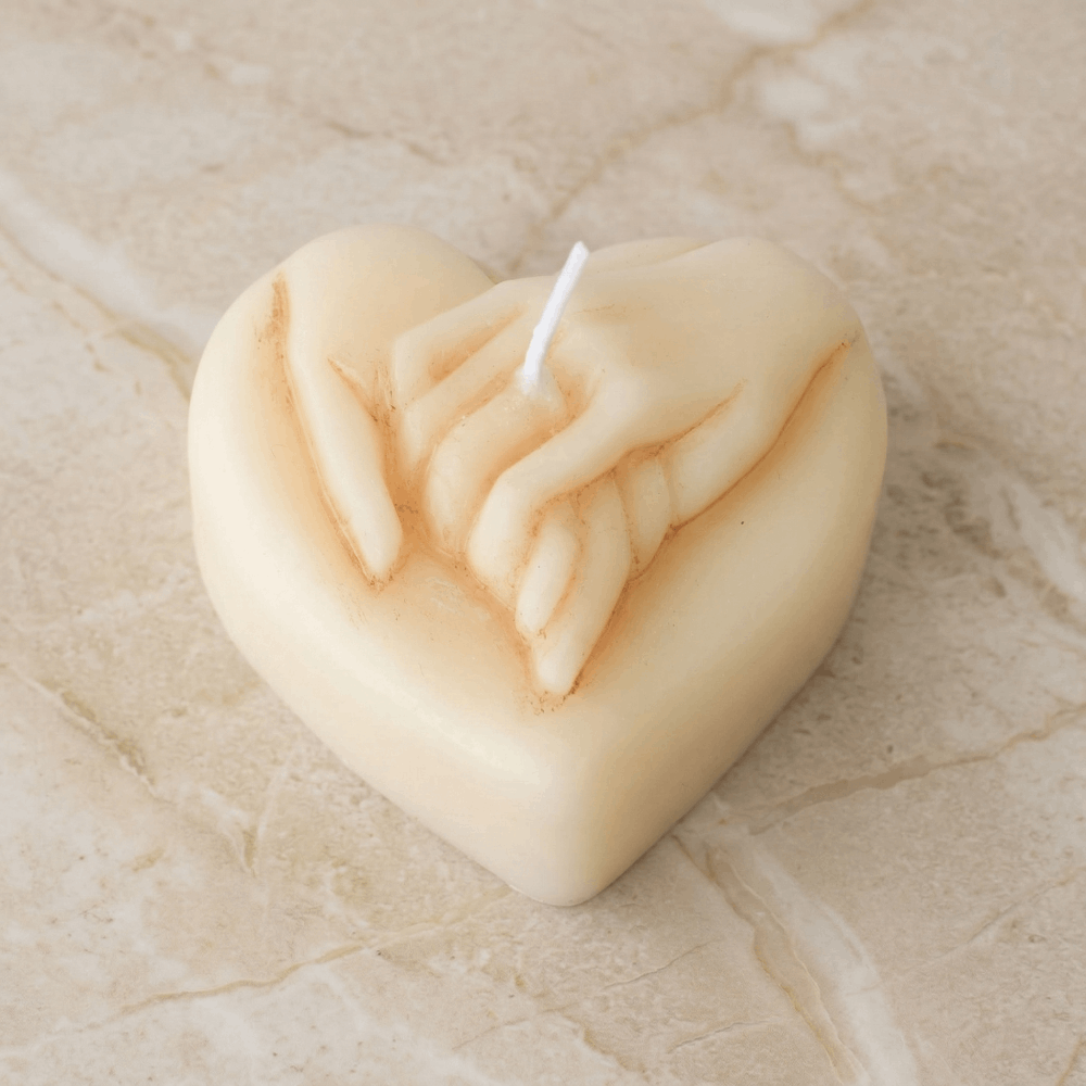 Vela de cera con forma de corazón de color crema o marfil en primer plano, mostrando una escultura detallada de dos manos entrelazadas con pigmento marrón rojizo en los pliegues para definición.