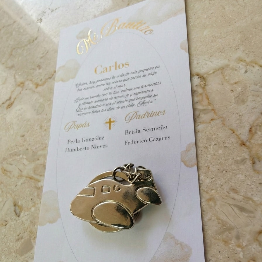 Tarjeta blanca para bolo de bautizo con impresión de letras doradas brillantes en foil, que incluye un llavero metálico dorado en forma de avioncito.